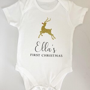 Puede incluir: Body de beb&eacute; blanco con un dise&ntilde;o de reno dorado con purpurina y el texto "Ella's FIRST CHRISTMAS". El reno tiene una nariz roja. El body tiene mangas cortas y corchetes en la parte inferior.