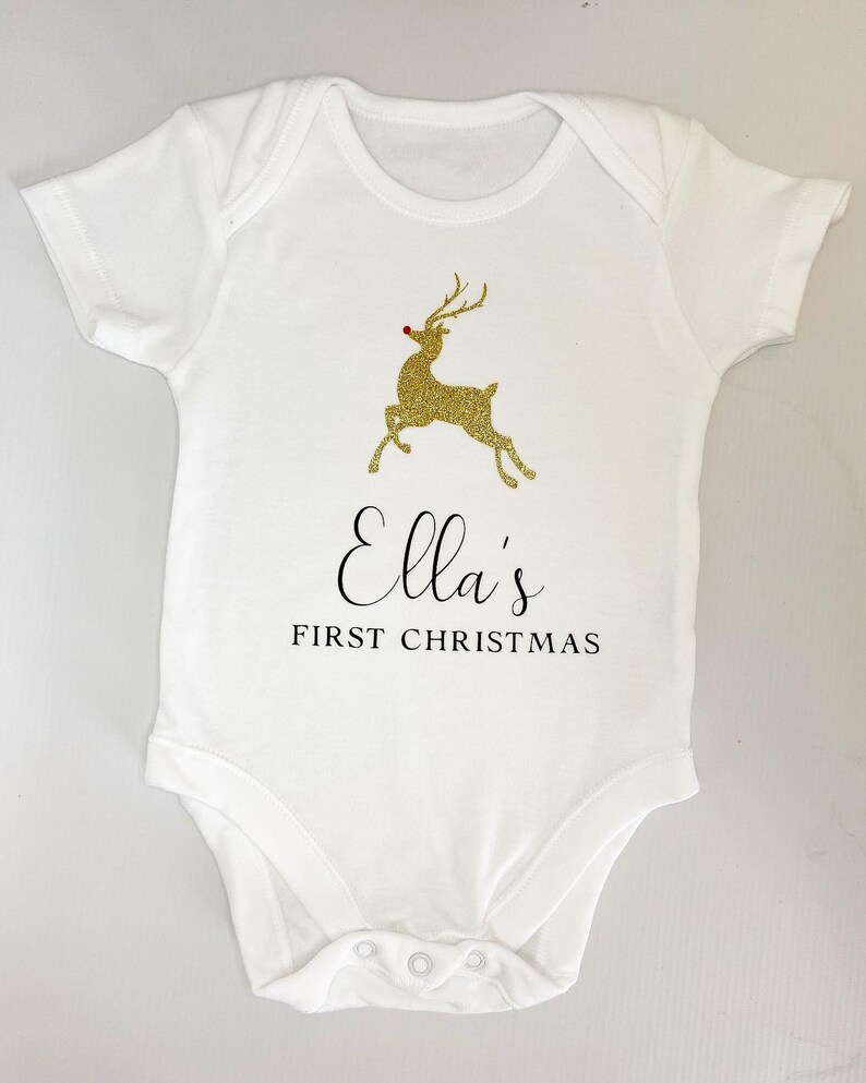 Puede incluir: Body de beb&eacute; blanco con un dise&ntilde;o de reno dorado con purpurina y el texto "Ella's FIRST CHRISTMAS". El reno tiene una nariz roja. El body tiene mangas cortas y corchetes en la parte inferior.