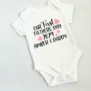 May include: White baby onesie with black text that reads "Our First Fathers Day 2024 Amber & Daddy" with pink hearts.