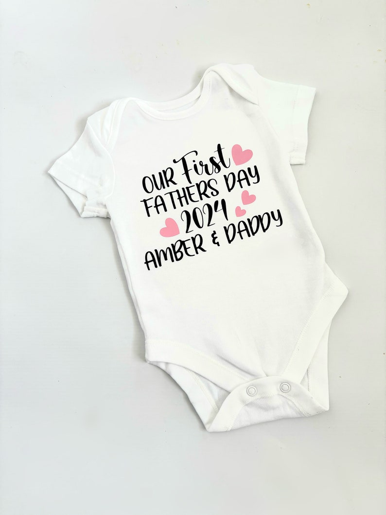 May include: White baby onesie with black text that reads "Our First Fathers Day 2024 Amber & Daddy" with pink hearts.