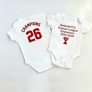 Op de afbeelding: Twee witte babyrompers. Eén heeft "CHAMPIONS 26" in rode letters op de rug. De andere heeft "Arsenal F.C. Premier League Champions 2025-2026" en een rode trofee.