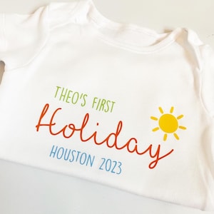 May include: A white short-sleeved baby bodysuit featuring the green text THEO'S FIRST, the orange script text Holiday, the blue text HOUSTON 2023, and a yellow sun graphic. The garment has a round neckline and short sleeves with ribbed trim.