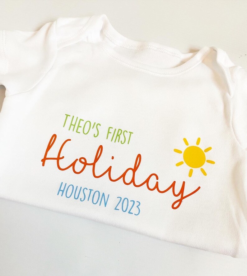 May include: A white short-sleeved baby bodysuit featuring the green text THEO'S FIRST, the orange script text Holiday, the blue text HOUSTON 2023, and a yellow sun graphic. The garment has a round neckline and short sleeves with ribbed trim.