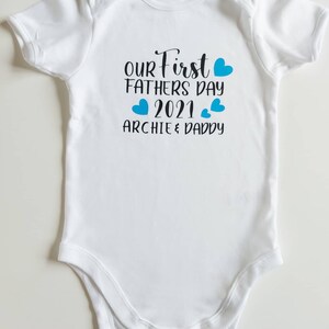 May include: White baby onesie with blue hearts and the text "Our First Fathers Day 2021 Archie & Daddy"