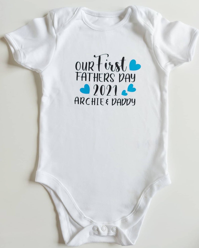 May include: White baby onesie with blue hearts and the text "Our First Fathers Day 2021 Archie & Daddy"
