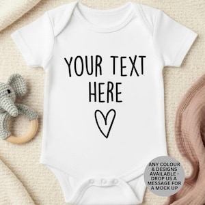 Könnte beinhalten: Ein weißer kurzärmeliger Babybody mit dem schwarzen Text "YOUR TEXT HERE" über einem handgezeichneten Herzsymbol. Das Kleidungsstück hat drei Druckknöpfe im Schritt und ein kreisförmiges graues Abzeichen mit Text zu individuellen Farb- und Designoptionen.