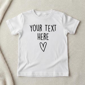 May include: A white short-sleeved t-shirt featuring the black text YOUR TEXT HERE printed in a simple sans-serif font, positioned above a hand-drawn black outline of a heart symbol centred on the front of the garment.