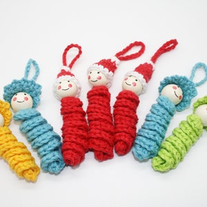 Könnte beinhalten: Eine Sammlung handgefertigter Häkelpuppen in verschiedenen Farben, darunter Gelb, Blau, Rot und Grün. Jede Puppe hat einen Holzkopf mit einem bemalten lächelnden Gesicht und eine Schlaufe zum Aufhängen. Die Puppen sind wahrscheinlich als Ornamente oder Dekorationsartikel gedacht.