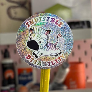 May include: A circular sticker with a shimmering, holographic background. It features a cartoon zebra with the words "INVISIBLE DISABILITY" in a circular arrangement. The zebra has black and white stripes.