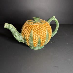 May include: A ceramic teapot shaped like a pineapple. The body is textured with a yellow and orange pattern, while the handle, spout, and lid are green. The teapot has a detailed design, with green leaves at the base.