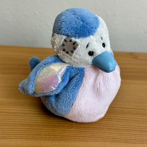 Pode incluir: Um pássaro de pelúcia com cabeça e asas azuis, barriga rosa macia e rosto branco com uma pequena mancha cinza. O pássaro tem um bico azul e detalhes em tecido iridescente e brilhante nas asas, com pequenos olhos pretos e detalhes costurados.