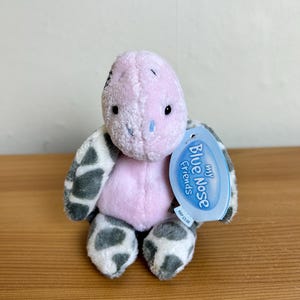 Könnte beinhalten: Ein Plüschtier mit hellrosa Kopf und Rumpf sowie grau-weiß gefleckten Gliedmaßen. Das Spielzeug hat eine kleine blaue Nase und schwarze Augen. Ein blaues Etikett an der Seite trägt die Aufschrift My Blue Nose Friends mit einem Preis von 5,00 £.