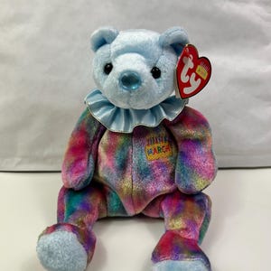 May include: A light blue plush bear with a ruffled collar and a multi-colored body. The bear has black eyes and a blue nose. A red heart-shaped tag is attached to the ear. The bear has a patch that says "March".