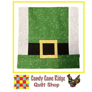 May include: Green fabric with white polka dots and a black belt with a yellow buckle. The fabric is sewn into a square shape. The text "Candy Cane Ridge Quilt Shop" is visible below the square.