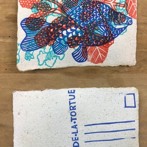 Puede incluir: Una postal blanca con un pez azul y rojo impreso con un dise&ntilde;o. La parte posterior de la postal tiene un sello azul y el texto "LE CRI DE LA TORTUE".