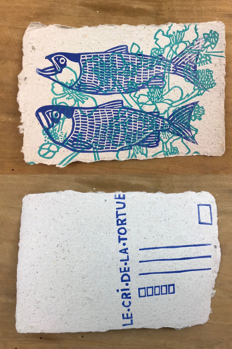 Puede incluir: Una postal con una impresi&oacute;n azul y verde de dos peces con dise&ntilde;os florales. La parte posterior de la postal tiene el texto "LE.CRI.DE.LA. TORTUE" impreso en azul.