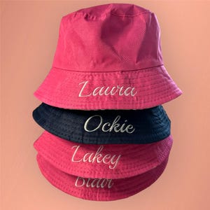 May include: Four pink and navy blue bucket hats stacked on top of each other. The hats are embroidered with the names Laura, Ockie, Lakey, and Blair in white thread.
