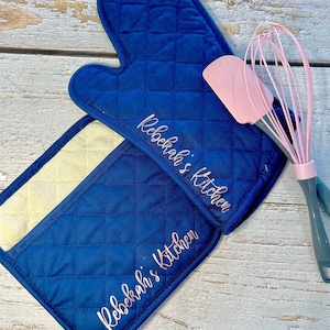 May include: A set of two blue quilted oven mitts with the text "Rebekah's Kitchen" embroidered in pink on each mitt. A pink silicone spatula and a pink whisk are also included.