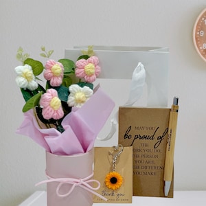 Pode incluir: Um conjunto de presentes com um buquê de flores de crochê rosa e branco em um cilindro rosa, um chaveiro de girassol, uma caneta de bambu, uma sacola de presente e um cartão com o texto "May you be proud of the work you do..."