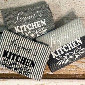 May include: Three kitchen towels with the text "[Name]'s Kitchen" in black and white lettering. The towels have different patterns, including a gray and white herringbone pattern, a black and white striped pattern, and a gray and white striped pattern.