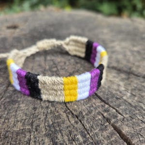 May include: A woven friendship bracelet featuring a repeating pattern of yellow, white, purple, black, and tan threads. The bracelet is constructed with a knotted design and includes adjustable braided ties at the ends for a secure fit.
