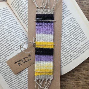 May include: A rectangular woven bookmark featuring horizontal stripes in beige, black, light purple, white, and yellow. The textile is mounted on a brown cardstock backing with a small tag labeled Emery Smith Studio and frayed fringe ends.