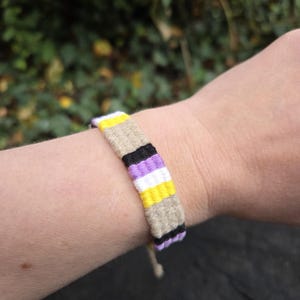 Op de afbeelding: Een geweven vriendschapsarmband met een herhalend patroon van horizontale strepen in geel, beige, zwart, wit en paars. De armband is gemaakt van meerdere draden die strak aan elkaar zijn geknoopt om een platte, duurzame band te vormen.