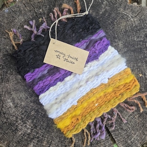 May include: A rectangular woven wall hanging featuring horizontal stripes of black, purple, white, and yellow yarn with frayed edges. A tan rectangular paper tag is attached with string, displaying the handwritten text emery smith studio.