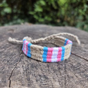 Puede incluir: Una pulsera tejida que presenta secciones alternas de fibra de color tostado natural y un patrón repetitivo de hilo azul brillante, blanco y rosa. La pulsera tiene largos lazos trenzados que se extienden a cada lado, hechos de la misma fibra.
