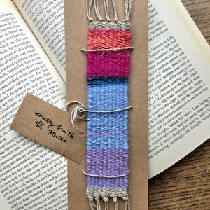 May include: A rectangular woven bookmark featuring a gradient of colors from red and pink at the top to blue and purple at the bottom, finished with beige fringe at both ends. A brown paper tag with the text Emery Smith Studio is attached.