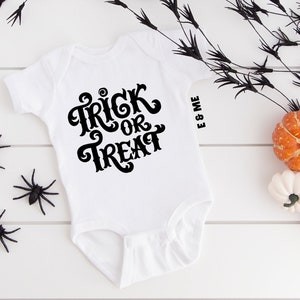 May include: White baby onesie with black text that says "Trick or Treat". The onesie is on a white wooden surface with black spider decorations and orange and white pumpkins.