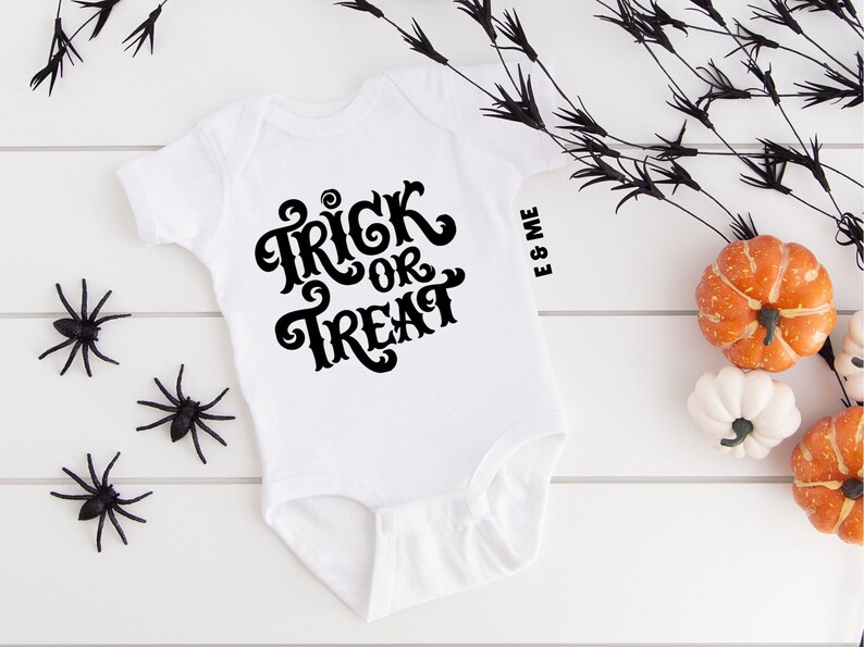 May include: White baby onesie with black text that says "Trick or Treat". The onesie is on a white wooden surface with black spider decorations and orange and white pumpkins.
