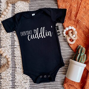 May include: Black baby onesie with white text that reads "Everyday I'm cuddlin". The onesie is on a white and gray striped surface with a wooden bead garland, a brown and white blanket, a wooden teether, and a small potted cactus.