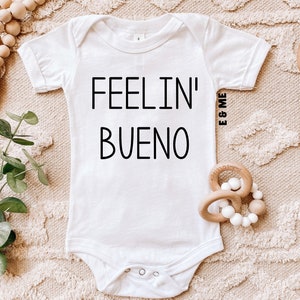 May include: White baby onesie with the phrase "FEELIN' BUENO" in black lettering. The onesie is short-sleeved and has snap closures at the bottom. A wooden teething ring and a beaded toy are next to the onesie on a textured surface.