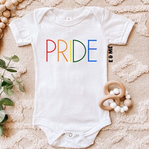 Pode incluir: Macac&atilde;o branco para beb&ecirc; com a palavra "PRIDE" em cores do arco-&iacute;ris. O texto "& EU" est&aacute; escrito em preto abaixo da palavra "PRIDE".