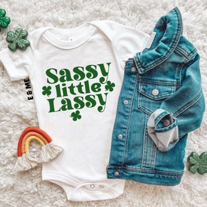May include: White baby onesie with green shamrock accents and the text "Sassy little Lass" in a retro font. The onesie is laid out on a white fluffy surface with a denim jacket and a rainbow-colored yarn rainbow.