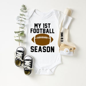 Może przedstawiać: Biały śpioszek dla niemowlęcia z czarnym napisem "My 1st Football Season" i brązową grafiką piłki nożnej.