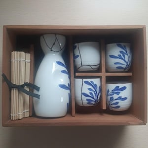 May include: A sake set in a wooden box. The set includes a white ceramic sake bottle with blue leaf designs, four matching cups, and bamboo skewers. The sake bottle has a black line design. The cups and bottle have blue leaf designs.