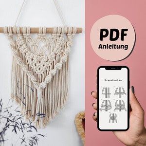 Puede incluir: Un tapiz de macramé blanco con una barra de madera en la parte superior. El tapiz tiene forma de lágrima y tiene un borla en la parte inferior. Un teléfono inteligente muestra una guía de instrucciones PDF para hacer el tapiz de pared, con el texto "PDF Anleitung" y "Kreuzknoten" visible.