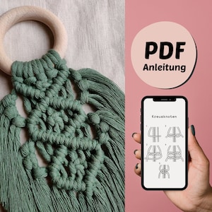 Może przedstawiać: Zielony wiszący makramowy z drewnianym pierścieniem na górze. Wisiorek wykonany jest z wzoru węzłów i ma długie frędzle na dole. Ekran telefonu pokazuje instrukcję PDF do wykonania wiszącego makramowego.
