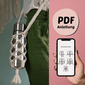 Puede incluir: Un porta botellas de macramé blanco con una termo de plata en su interior. El porta botellas tiene un borla en la parte inferior. La pantalla de un teléfono muestra instrucciones para hacer un nudo de macramé. El texto en la pantalla dice "Kreuzknoten". El teléfono está sostenido por una mano. Un círculo con el texto "PDF Anleitung" está en el fondo.