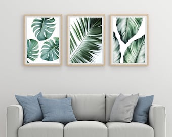 May include: Three framed prints of tropical leaves in shades of green against a white background. The prints are hanging on a white wall above a light gray sofa with blue throw pillows.