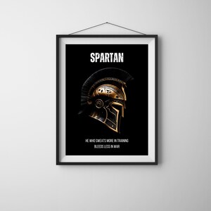 May include: Framed art print featuring a Spartan helmet design. The helmet is gold and black, with the word "SPARTAN" at the top and the quote "HE WHO SWEATS MORE IN TRAINING BLEEDS LESS IN WAR" at the bottom. The background is black.