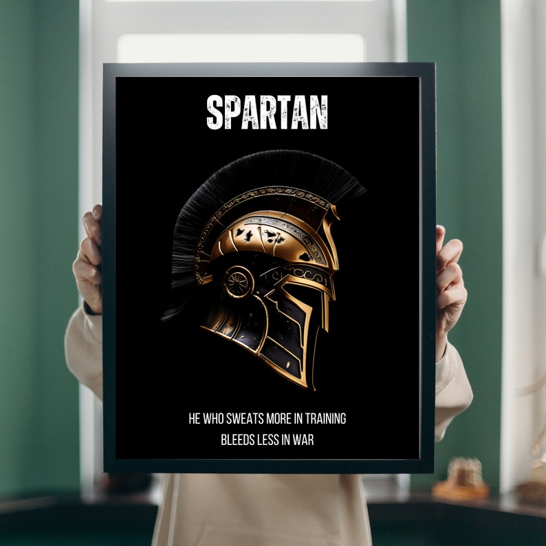 May include: Black framed print featuring a gold and black Spartan helmet design. The word "SPARTAN" is displayed at the top, with the quote "HE WHO SWEATS MORE IN TRAINING BLEEDS LESS IN WAR" below the helmet.