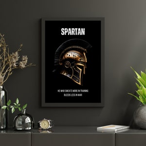 May include: Black framed wall art featuring a gold and black Spartan helmet on a black background. The word "SPARTAN" is at the top, with the quote "HE WHO SWEATS MORE IN TRAINING BLEEDS LESS IN WAR" below the helmet.