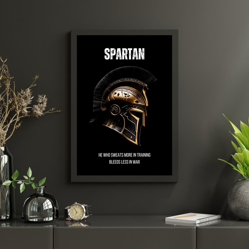 May include: Black framed wall art featuring a gold and black Spartan helmet on a black background. The word "SPARTAN" is at the top, with the quote "HE WHO SWEATS MORE IN TRAINING BLEEDS LESS IN WAR" below the helmet.