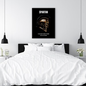 May include: A framed black poster with the word "SPARTAN" at the top and a gold and black Spartan helmet illustration. The text "HE WHO SWEATS MORE IN TRAINING BLEEDS LESS IN WAR" is below the helmet. The poster is displayed above a bed.