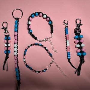 May include: A selection of beaded accessories, including keyrings and bracelets, in the transgender pride flag colours: pink, white, and blue. The beads are strung on black cord and have silver-tone clasps.