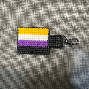 May include: A small crocheted keychain featuring the colors of the non-binary pride flag: yellow, white, purple, and black. The keychain has a black metal ring.