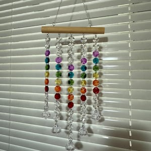 May include: A rainbow crystal suncatcher with a wooden dowel and clear crystal beads hanging from a silver chain. The suncatcher is hanging in front of a white window blind.